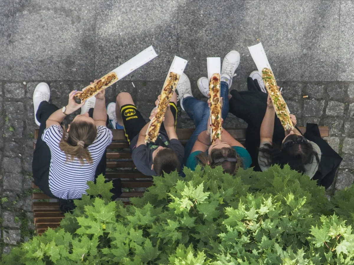 Four people on a bench eating long slices of pizza beneath a leafy tree seen from above